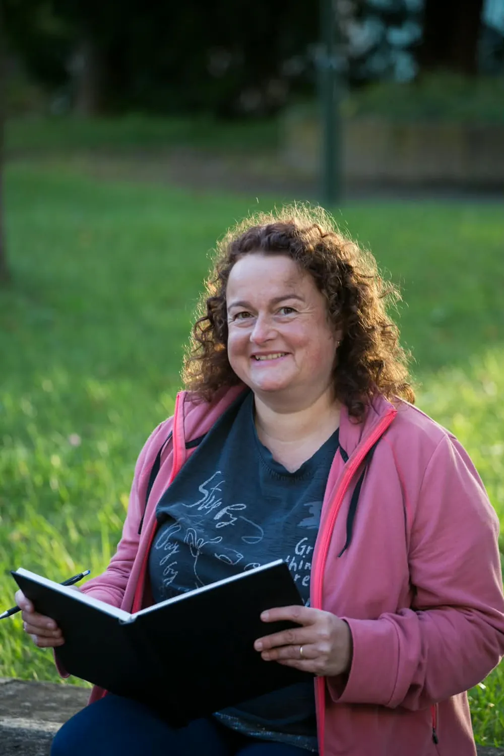 Christiane Wenzel sitzt lächelnd auf einer Wiese, ein aufgeschlagenes Notizbuch und einen Stift in den Händen.