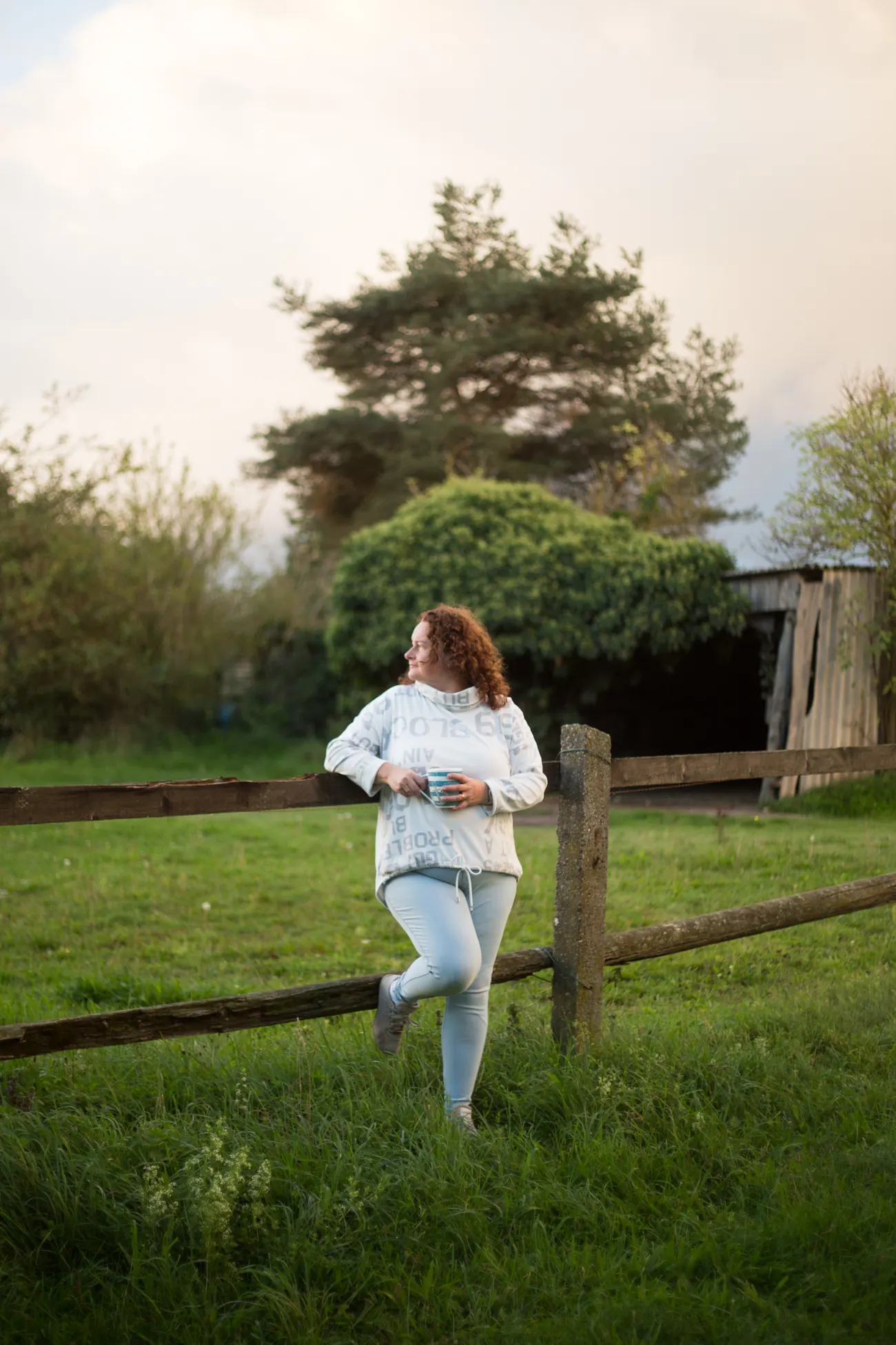 Christiane Wenzel lehnt an einem Holzzaun auf einer Wiese, eine Tasse in der Hand, und blickt in die Abendsonne.