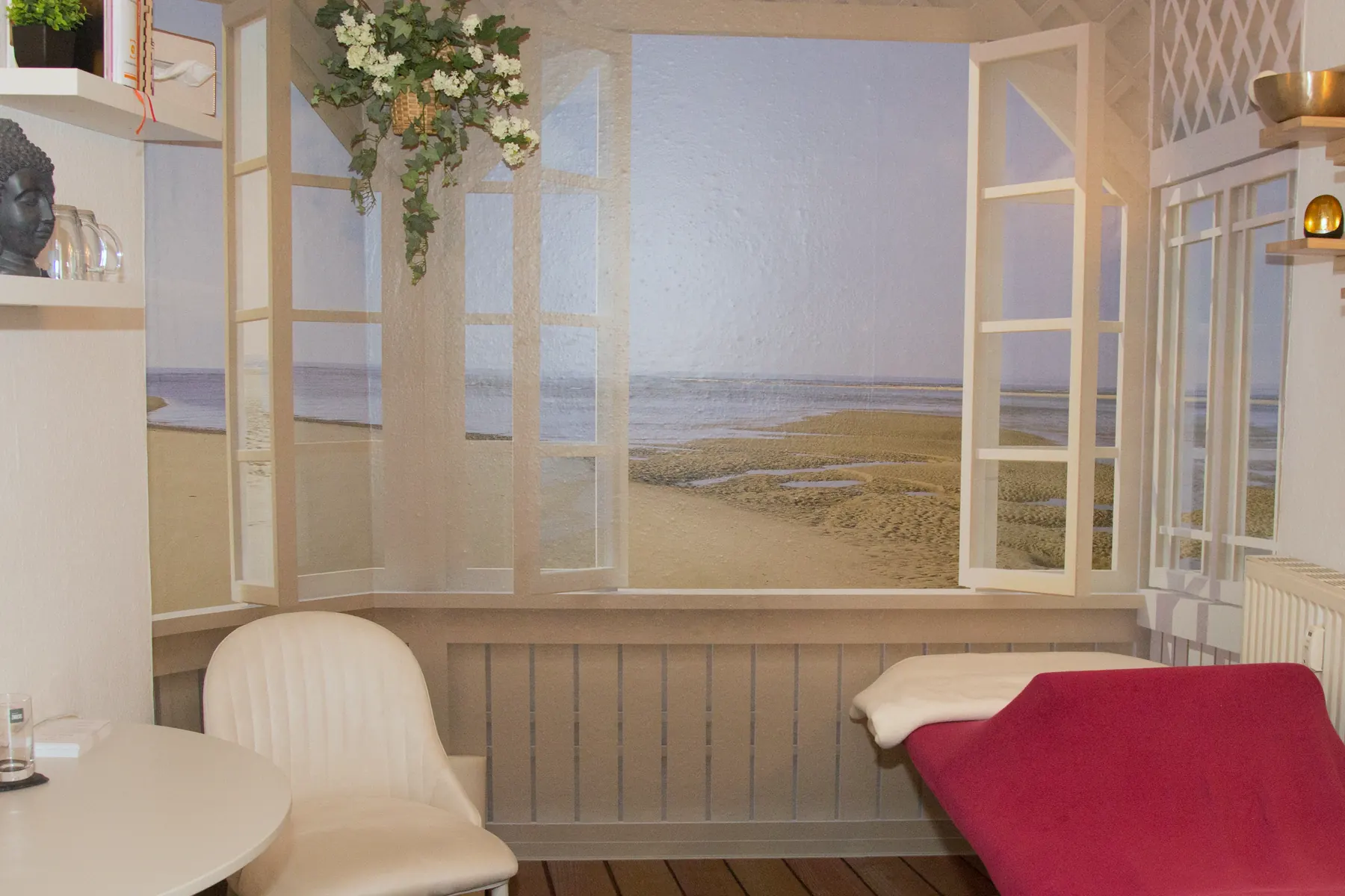 Christianes Raum in Lobstädt: eine Liege mit roter Decke, ein runder Tisch mit Stuhl und eine Fenstertapete mit Blick auf Strand und Meer.